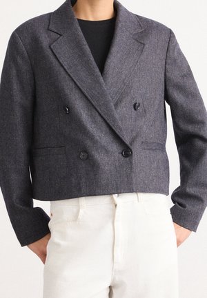 Osoba nosząca krótki ciemnoszary dwurzędowy blezer z szerokimi klapami na czarnej bluzce i białych spodniach z wysokim stanem, ręce w kieszeniach.