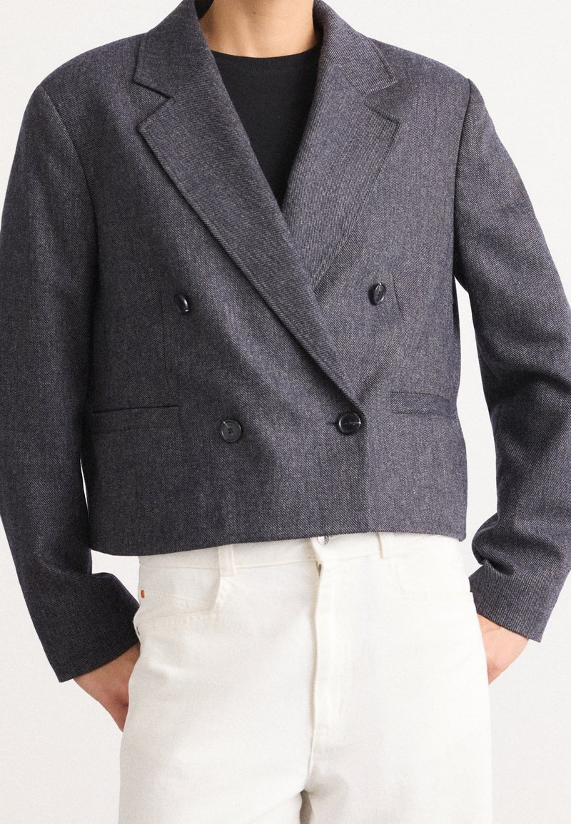 Personne portant un blazer court gris foncé à double boutonnage avec de larges revers sur un top noir et un pantalon blanc taille haute, mains dans les poches.