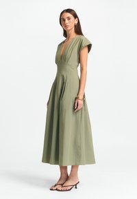 Tamsiai žalios midi suknelė su gilia iškirpte, trumpomis rankovėmis ir A formos sijonu. Tekstūruotas audinys su subtiliais raukiniais, derinamas su minimalistiniais sandalais.