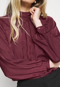Blusa bordeaux a maniche lunghe con scollatura ruffle, dettagli plissettati e polsini con bottoni. Tessuto morbido e leggero con un design fluido.