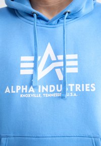 Šviesiai mėlyna medvilnės mišinio bliuzonas su baltu logotipu ir užrašu "ALPHA INDUSTRIES", su raišteliais ir priekiu kišene.
