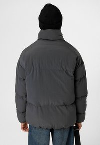 Donsjack in donkergrijze stof met een hoge kraag, elastische manchetten en puffy secties. Gekozen over een lichtblauwe spijkerbroek en een zwarte beanie.