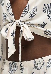 Gros plan d'un torse à la peau foncée portant un tissu blanc à imprimé botanique noir, noué en un nœud au niveau de la taille, laissant voir le nombril et la ceinture élastique.