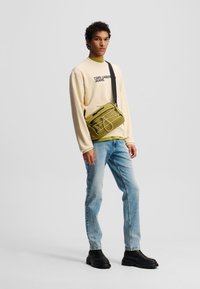 Hanorac crem cu logo, suprapus peste o cămașă verde. Blugi albaștri, cizme negre masive și geantă crossbody verde oliv cu detalii cu fermoar.