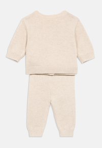 Bézs kötött babaruha rövid ujjú pulóverrel és hozzá illő nadrággal, bordázott mandzsettákkal és szegéllyel, fehér háttéren laposan elhelyezve.