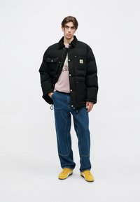 Chaqueta de plumas negra con cuello, que presenta botones a presión y un bolsillo en el pecho. Usada sobre una camiseta rosa, combinada con jeans azules holgados y zapatos amarillos.