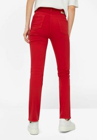 Jeans skinny rossi con una texture liscia, caratterizzati da una vita media, due tasche posteriori e un'etichetta logo sulla vita. Indossati con sneaker bianche.