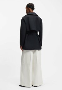 Czarna krótka kurtka z szerokimi klapami i przewiązanym paskiem, zestawiona z luźnymi białymi spodniami. Gładka tkanina i czyste linie.