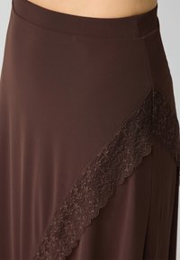 Bruine rok gemaakt van een gladde stof met een kanten randdetail aan één zijde, kenmerkend door een subtiele glans en elegante val.