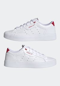 Vita lädersneakers med perforerat blommönster, röda accenter vid hälen och varumärkeslogotypen, platt vit gummisula och standard snörningsdesign.