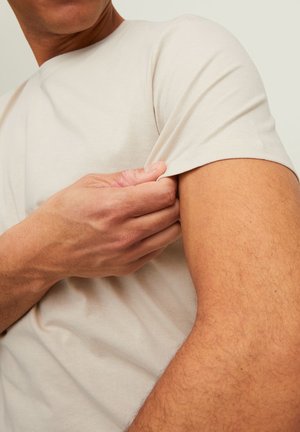 Homme tenant et étirant la manche courte beige d'un t-shirt sur son bras supérieur nu, sur un fond neutre.