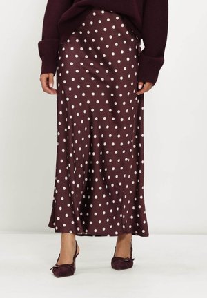 Vrouw draagt een lange bruine rok met witte stippen en bijpassende bruine slingback hakken, staat op een witte vloer.
