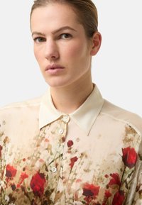 Durchscheinendes Blusenhemd mit Blumenmuster, ausgestattet mit einem weichen cremefarbenen Kragen und einem Aquarellmuster in Rot- und Grüntönen, das eine zarte Textur erzeugt.