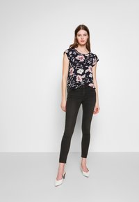 Top preto com padrão floral e mangas curtas, combinado com jeans pretos justos e saltos brancos de bico fino. Tecido suave, ajuste confortável.
