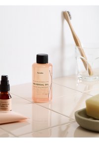Pink plastic bottle of "Universal Oil" with black cap, featuring label. Nearby are a dark glass dropper bottle and a bar of soap on beige tiles.