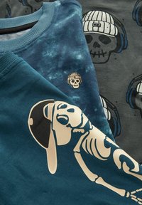 Tres camisetas exhibidas: una de color turquesa con un pequeño logo de calavera, una de color gris oscuro con un gráfico de calavera, y una de color azul claro con un diseño de esqueleto.