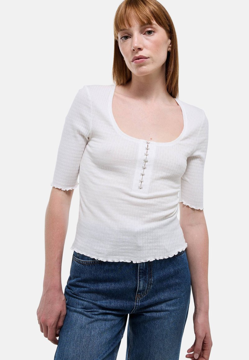 Crop top blanc à côtes avec des manches courtes, décolleté en forme de goutte et fermeture décorative par boutons à l'avant. Présente un ourlet ondulé.