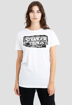 Junge Frau, die ein weißes T-Shirt mit einem schwarzen, abgenutzten "Stranger Things"-Logo trägt, steht vor einem schlichten, hellgrauen Hintergrund.