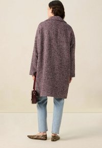 Manteau en chevrons long bordeaux et crème, coupe ample, avec un large col. Un jean bleu et des ballerines à motifs foncés complètent le look.