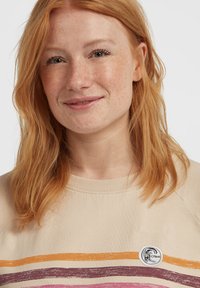 Red-haired woman with freckles wearing a beige sweater with orange, brown, and pink stripes and an O’Neill logo patch, smiling gently.