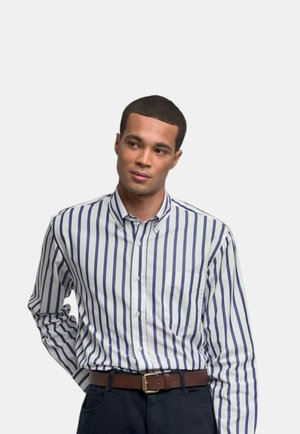 Uomo con una camicia bianca a righe verticali blu scuro e pantaloni scuri, in piedi con una mano dietro la schiena.