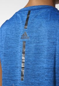 Zadní pohled na osobu nosící modré sportovní tričko Adidas s černými vertikálními reflexními pruhy a logem Adidas na horní části zad.