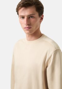 Béžová mikina s dlouhým rukávem s klasickým kulatým výstřihem, z měkké tkaniny, hladké textury a minimalistického designu bez viditelných log nebo vzorů.