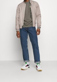 Chaqueta bomber marrón, sudadera oliva con logo, pantalones vaqueros rectos azules, zapatillas blancas con detalles multicolores. Conjunto casual exhibido.