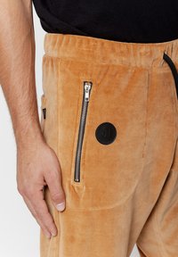 Welurowe dżinsy w ciepłym beżowym odcieniu z bocznymi kieszeniami na zamek, elastycznym paskiem w talii i okrągłą czarną łatą z logo na udzie.