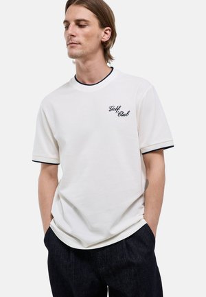 Jeune homme aux cheveux mi-longs portant un t-shirt blanc à manches courtes avec le texte « Golf Club » et un pantalon foncé, les mains dans les poches, regardant en bas à gauche.