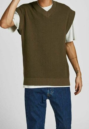 Mand iført olivengrøn ærmeløs strikket vest over hvid t-shirt og blå jeans, med synlig tatovering og armbånd på højre arm.