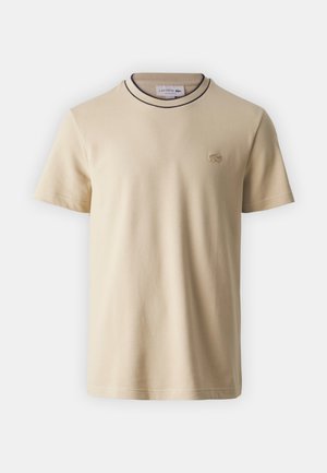 Camiseta de manga corta color beige, hecha de algodón, con un cuello de contraste y un pequeño logo bordado en el pecho. Textura suave.