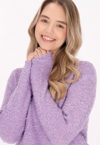 Pulover texturat mov, cu mâneci lungi; material moale, pufoase; purtătorul își strânge mâinile sub bărbie, fundal deschis.