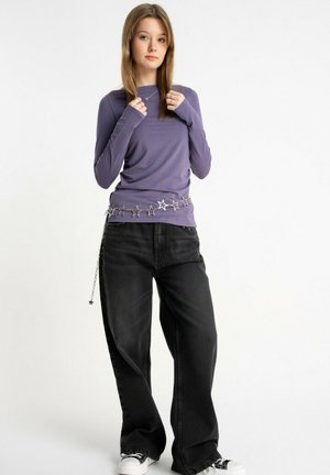 Młoda kobieta ubrana w fioletową koszulę z długim rękawem, czarne szerokie spodnie dżinsowe, czarno-białe trampki oraz pasek w kształcie łańcucha z gwiazdkami, stoi na białym tle.