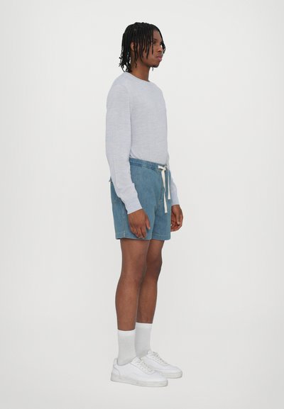 Chemise à manches longues gris clair, shorts en denim bleu avec taille élastique et cordon de serrage, chaussettes blanches et baskets blanches.