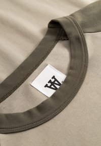 Beež puuvillast T-särk, millel oliivalroheline ümarsärk. Nähtav silt musta logoga. Sile tekstuur ja lihtne disain.