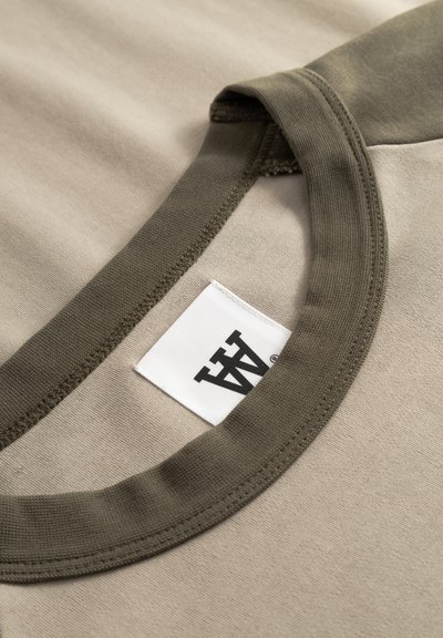 Μπεζ βαμβακερό T-shirt με λαιμόκοψη σε ελιά πράσινο. Ορατή ετικέτα με μαύρο λογότυπο. Ομαλή υφή με απλό σχέδιο.