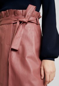 Vaaleanpunainen nahkahame, jossa on röyhelövyötärö ja solmittu vyö. Sileä pinta, jossa näkyvät saumat. Mukana tummansiniset ribbitekniikalla valmistetut hihat.