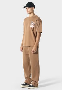 Ruskea ylipitkä t-paita, jossa on vaaleanpunainen lehtikuvio taskussa, yhdistettynä vastaaviin ruskeisiin verkkareihin ja mustiin lenkkareihin, joissa on valkoiset pohjat.