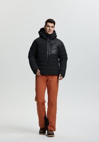 Chaqueta de plumas negra con capucha y media cremallera, combinada con pantalones impermeables naranjas y botas marrones. Acentos negros notables en los bolsillos.