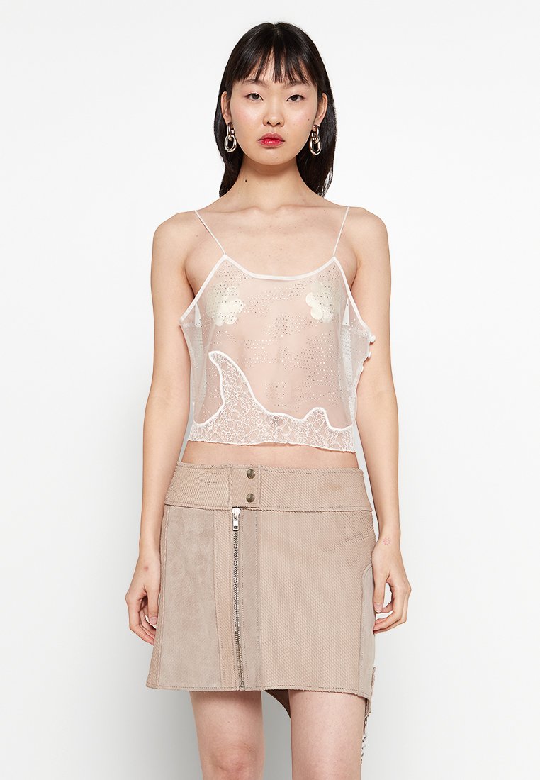 Donna che indossa una camicia in pizzo bianco trasparente con applicazioni floreali e una mini gonna beige con texture, dotata di zip frontale e bottoni, in piedi su uno sfondo bianco.