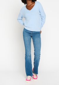 Pull bleu clair à col en V, douillet, associé à un jean évasé. Les chaussures présentent des touches roses. Tenue décontractée sur un fond neutre.