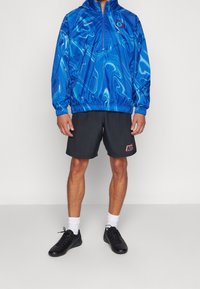Mężczyzna w niebieskiej kurtce z wzorem Chelsea FC z półzamkiem, czarnych szortach Nike, białych skarpetach i czarnych butach sportowych, stojący na tle białym.