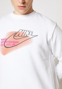 Vit sweatshirts med rund halsringning som har en framträdande svart "NIKE" logotyp, en orange gradient swoosh och rosa text som säger "NIKE SPORTSWEAR EST 1972."