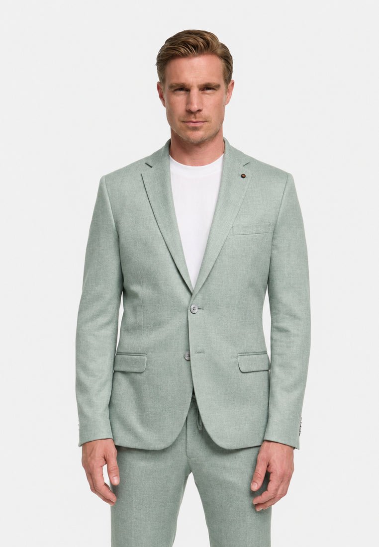 Hombre con cabello rubio corto que lleva una chaqueta de traje gris claro y pantalones a juego sobre una camiseta blanca de cuello redondo, mirando hacia adelante.
