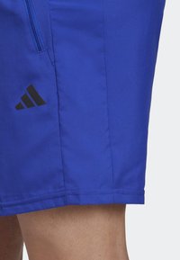 Blå sportshorts med svart Adidas-logga på vänster sida och en synlig dragkedjelomme ovanför loggan, bärs på nakna ben.