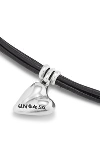 Pulsera de cuero negro con un brillante charm en forma de corazón plateado, grabado con "UNO de 50," sujeto con anillos plateados.