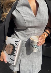 Harmaa, hihaton mekko, jossa on V-artikkeli ja tekstuurinen kangas. Pitelee lattejuomaa brändätyssä kupeissa ja aikakauslehteä, jossa on naisen muotokuva.