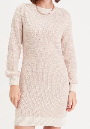 Kobieta ubrana w jasnobeżową dzianinową sukienkę z długim rękawem, nosząca naszyjnik z łańcuszka, stojąca na białym tle.