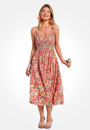 Młoda kobieta w różowej sukience midi w kwiaty i sandałach, uśmiechająca się, trzymająca rożek lodów z kwiatowymi ozdobami we włosach i biżuterią.